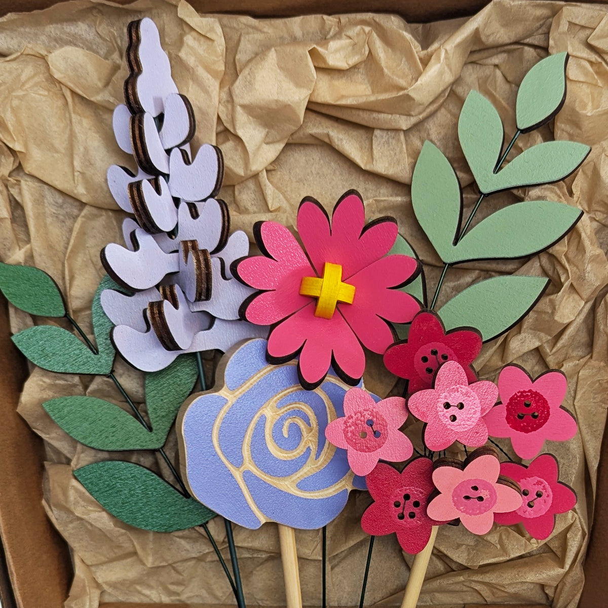 Signature Blooms in Macaron, a close crop of all four wooden blooms and two foliage stems in bright pink and violet, arranged in the box against brown tissue