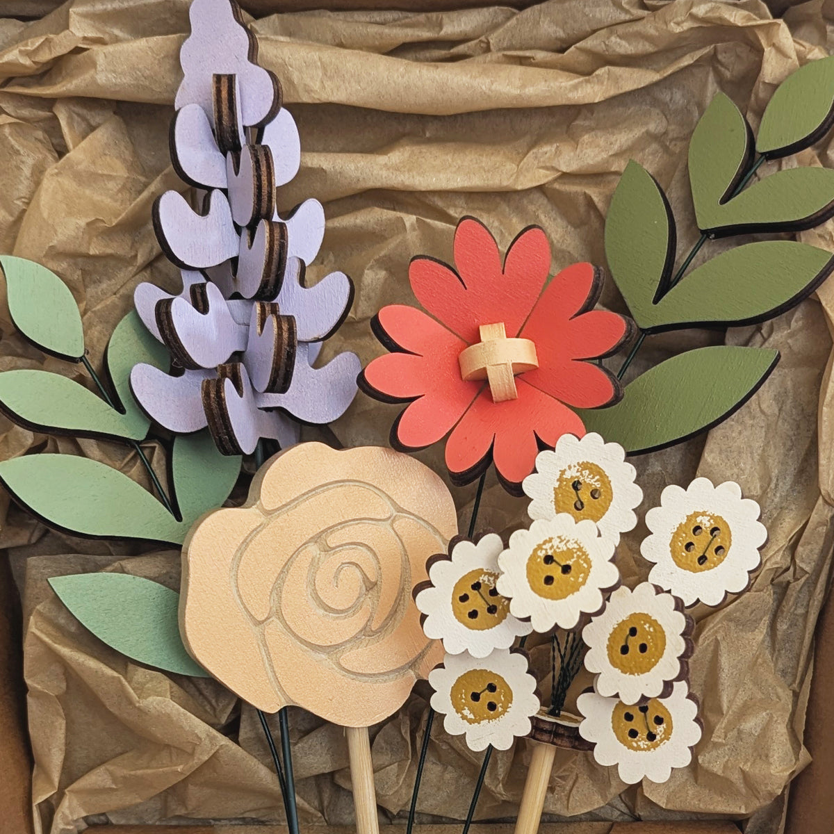 Signature Blooms in Sorbet, a close crop of all four wooden blooms and two foliage stems in muted mustard, lilac, peach, coral and sage, arranged in the box against brown tissue