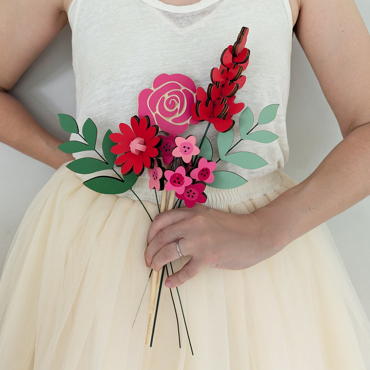 Signature Blooms in Framboise, four wooden blooms and two foliage stems in red, fuchsia and pink, held at waist height against pale neutral clothing
