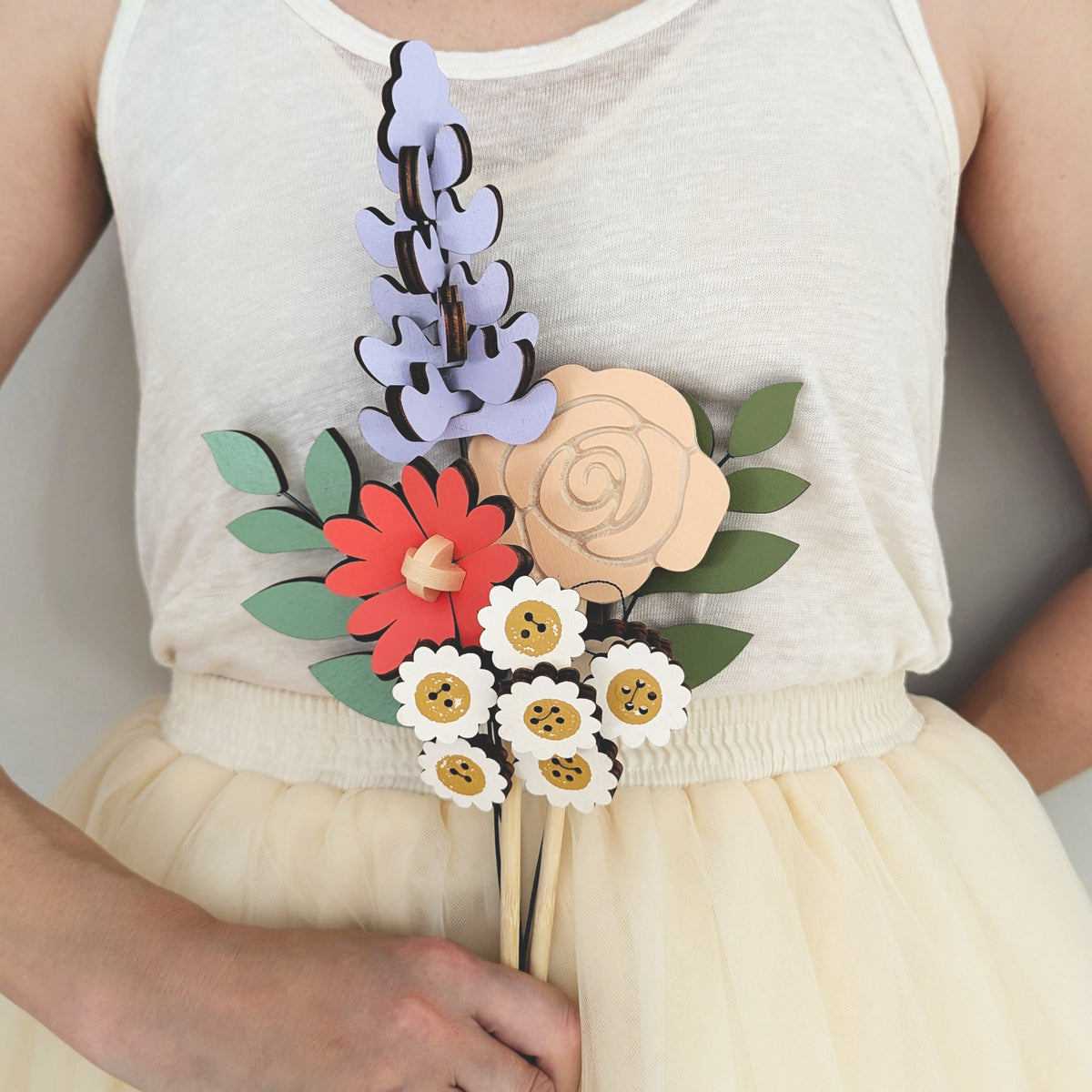 Signature Blooms in Sorbet, four wooden blooms and two foliage stems in muted mustard, lilac, peach, coral and sage, held at waist height against pale neutral clothing