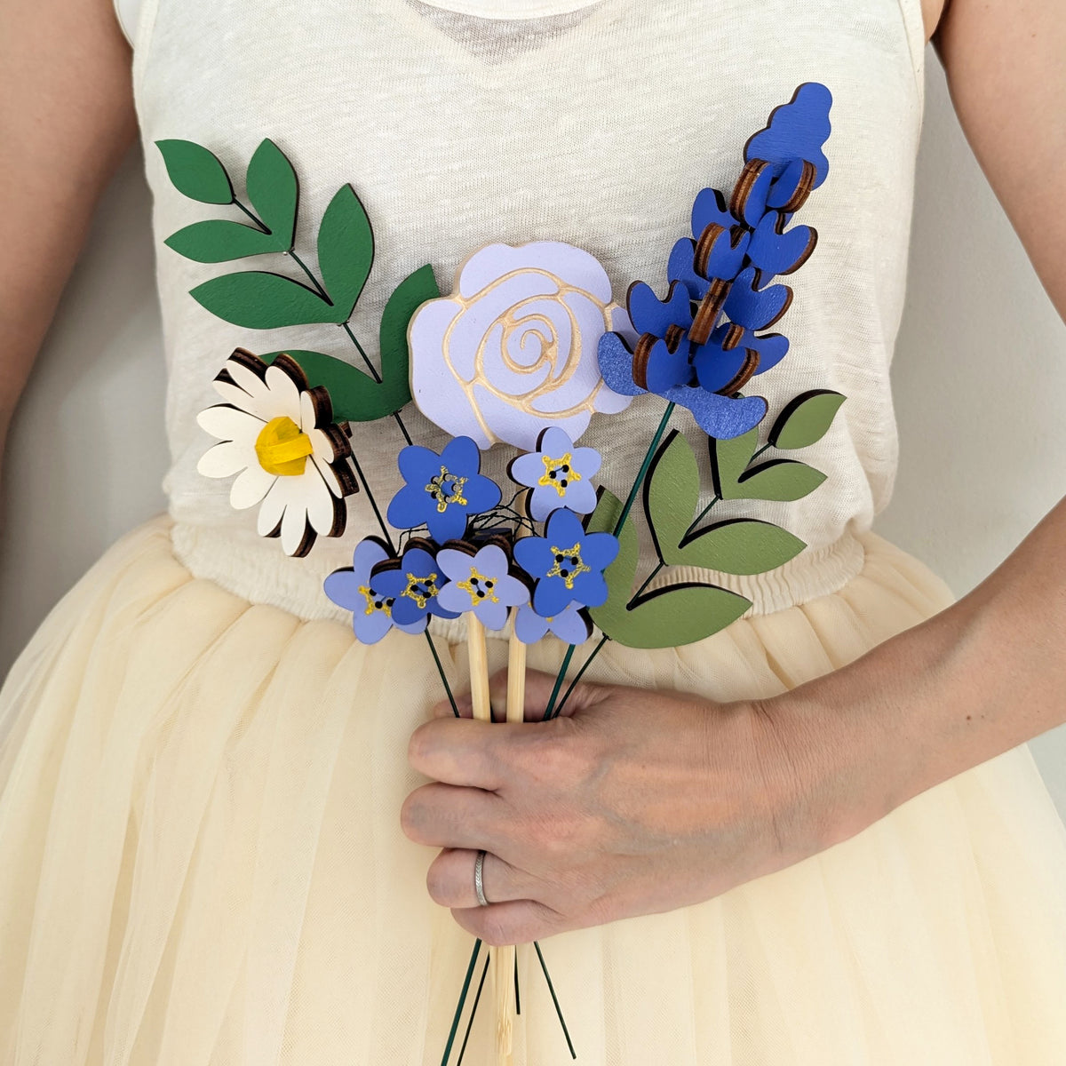 Signature Blooms in Parma Violet, four wooden blooms and two foliage stems in violet and lilac, held at waist height against pale neutral clothing