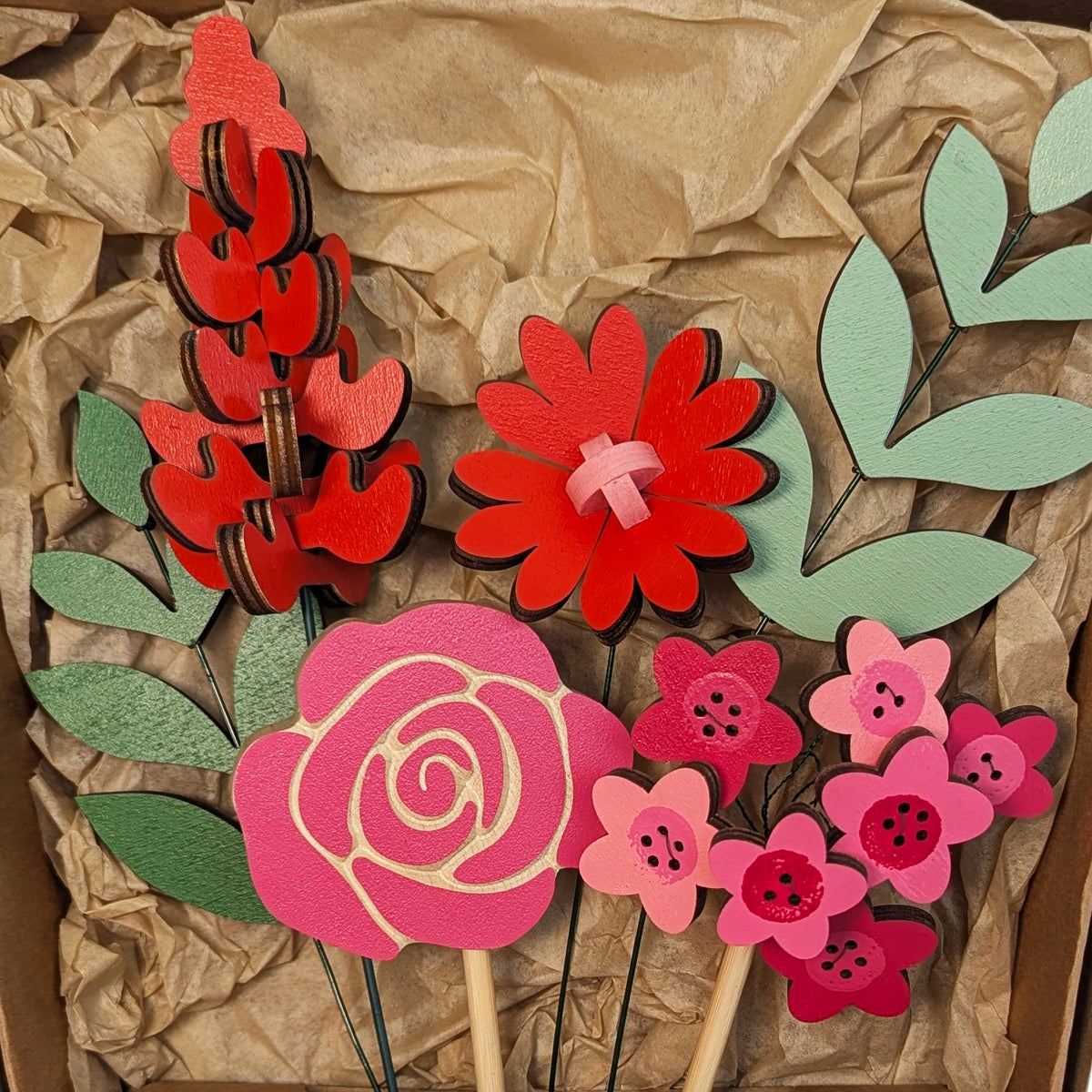 Signature Blooms in Framboise, a close crop of all four wooden blooms and two foliage stems in red, fuchsia and pink, arranged in the box against brown tissue 
