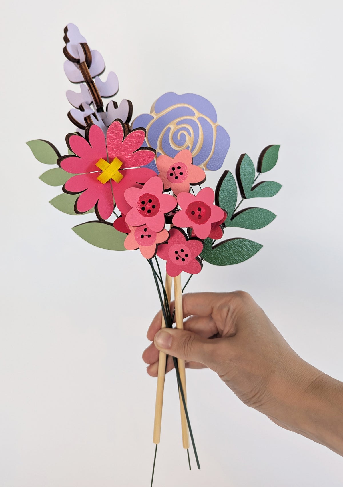 Signature Blooms in Macaron, four wooden blooms and two foliage stems in bright pink and violet, held in one hand at a second angle against a plain white background 