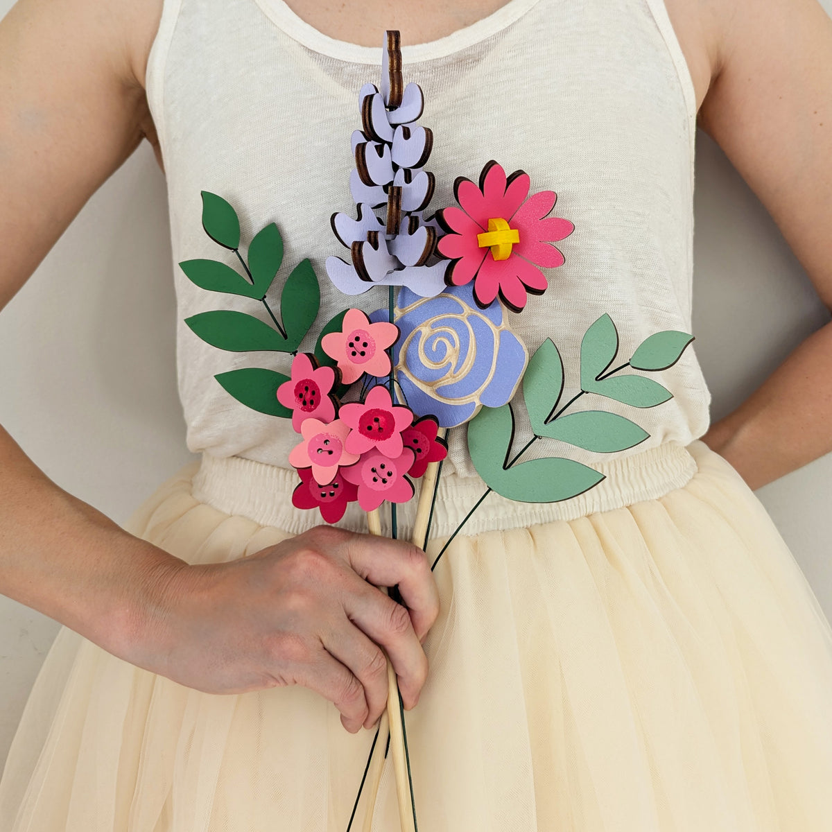 Signature Blooms in Macaron, four wooden blooms and two foliage stems in bright pink and violet, held at waist height against pale neutral clothing 
