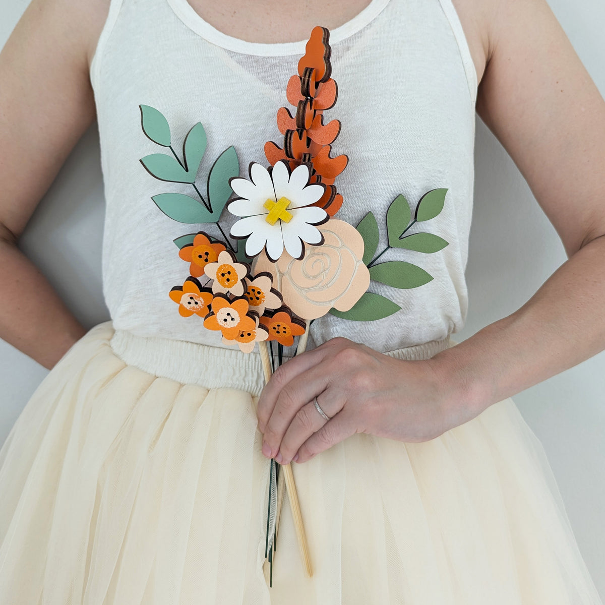 Signature Blooms in Satsuma, four wooden blooms and two foliage stems in warm orange, held at waist height against pale neutral clothing 
