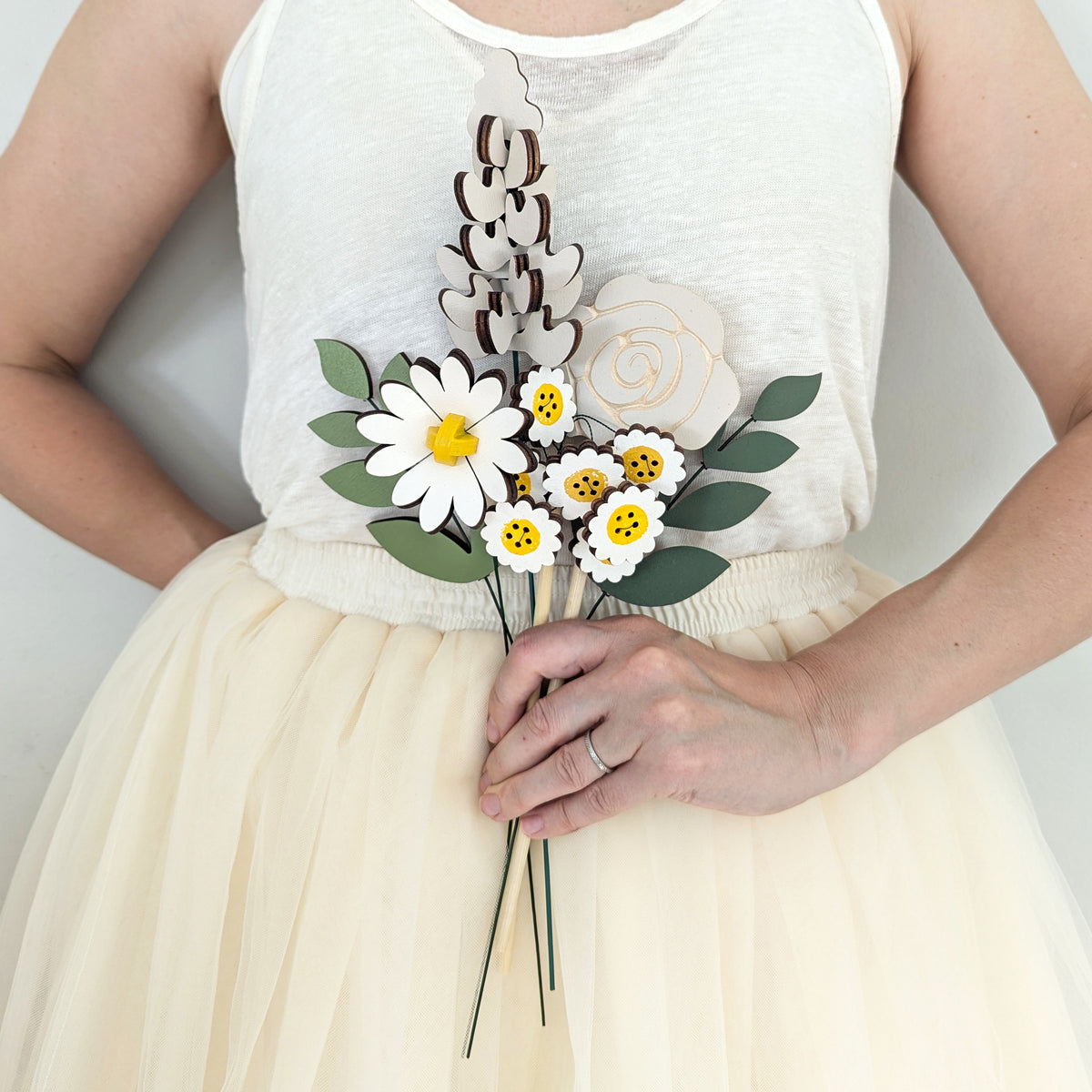 Signature Blooms in Sunny Side, four wooden blooms and two foliage stems in grey and white with a touch of sunshine yellow, held at waist height against pale neutral clothing 
